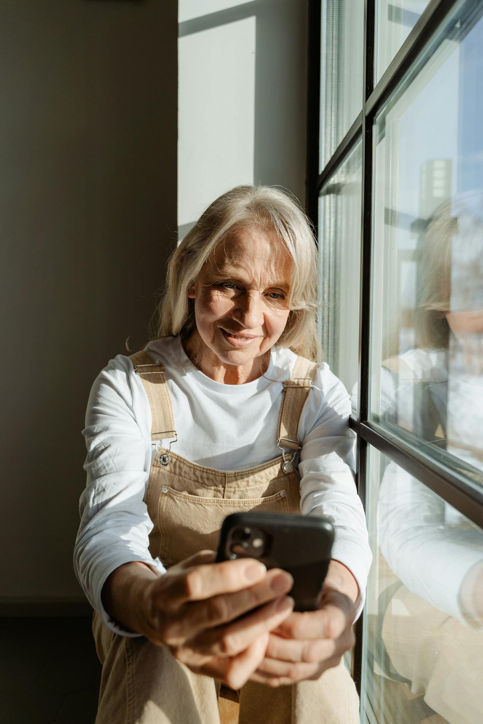 Senior woman relaxing at home, using smartphone by the window, enjoying leisure time.